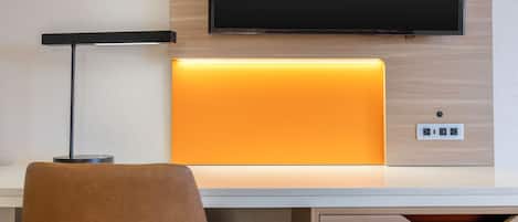 A hotel room with a flat-screen TV mounted on the wall, a desk with a lamp, and a chair.
