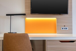 Habitación de hotel con televisión de pantalla plana montada en la pared, un escritorio con lámpara y una silla.