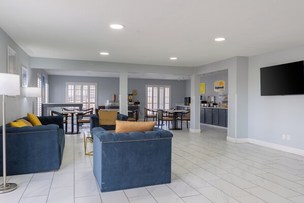A spacious living area with a dining table and chairs, a kitchen area with a flat-screen TV, and a large window with blinds.