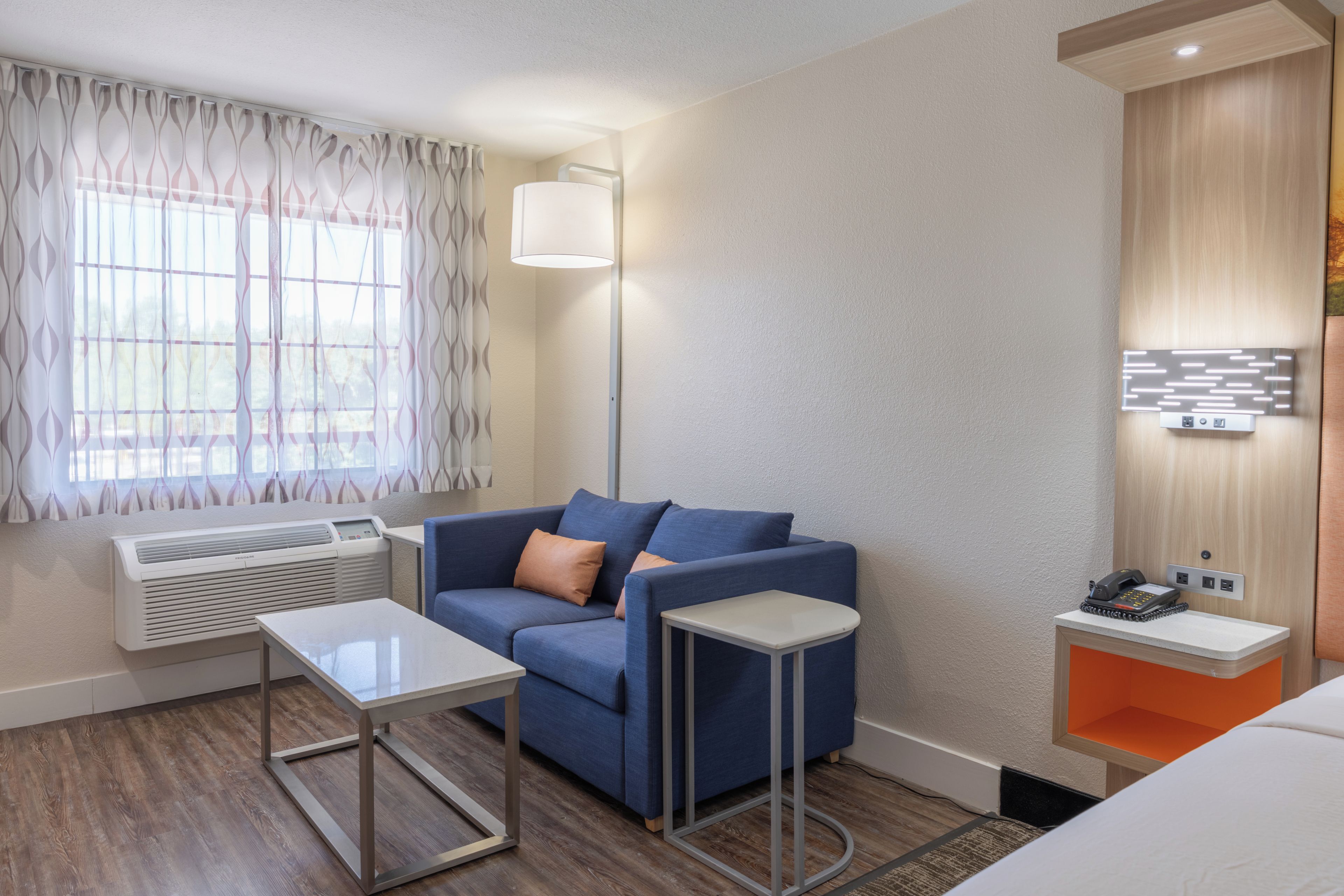 A hotel room with a blue sofa, a small table, a standing lamp, a heater, a phone, and a wall-mounted light fixture.