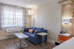 A hotel room with a blue sofa, a small table, a standing lamp, a heater, a phone, and a wall-mounted light fixture.