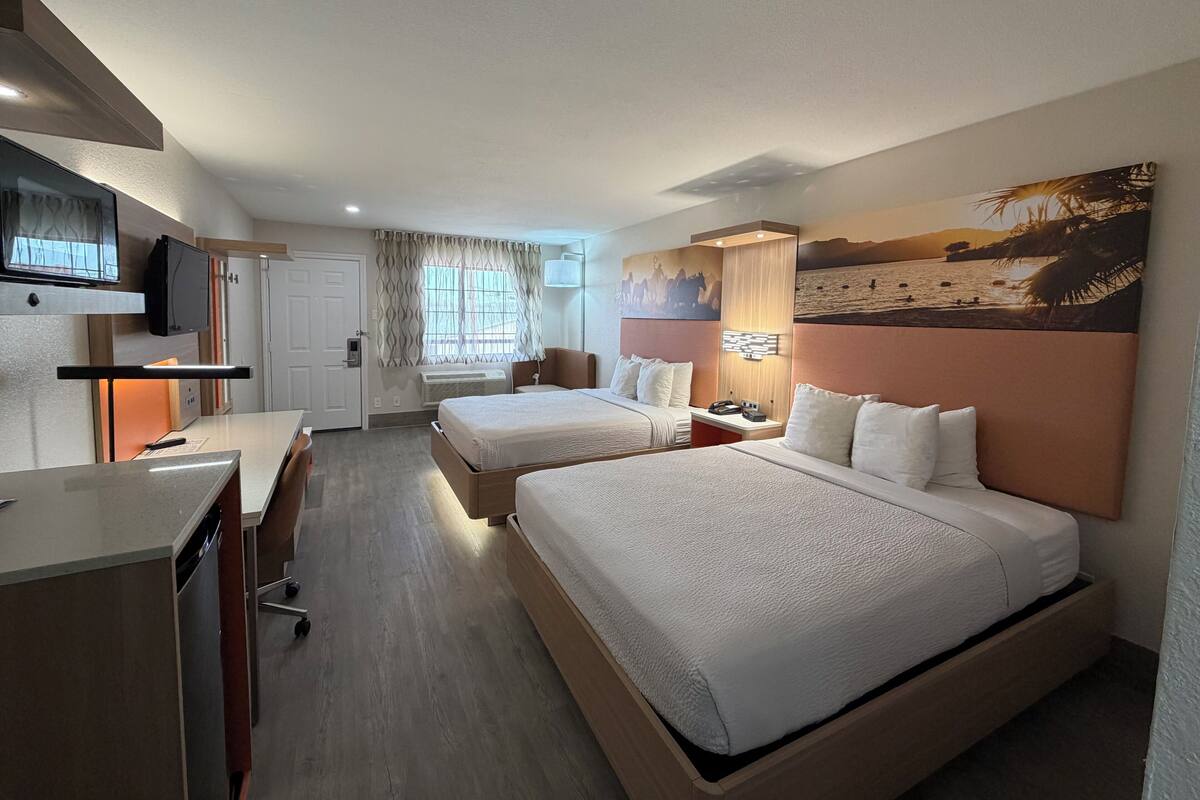 Ein Hotelzimmer mit zwei Betten, einem Schreibtisch, einem Fernseher und einem Bild mit einer Strandansicht.