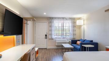 Habitación de hotel con cama, escritorio, silla, sofá, televisor, ventana con cortinas y una lámpara de pared.