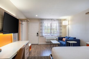 Habitación de hotel con cama, escritorio, silla, sofá, televisor, ventana con cortinas y una lámpara de pared.