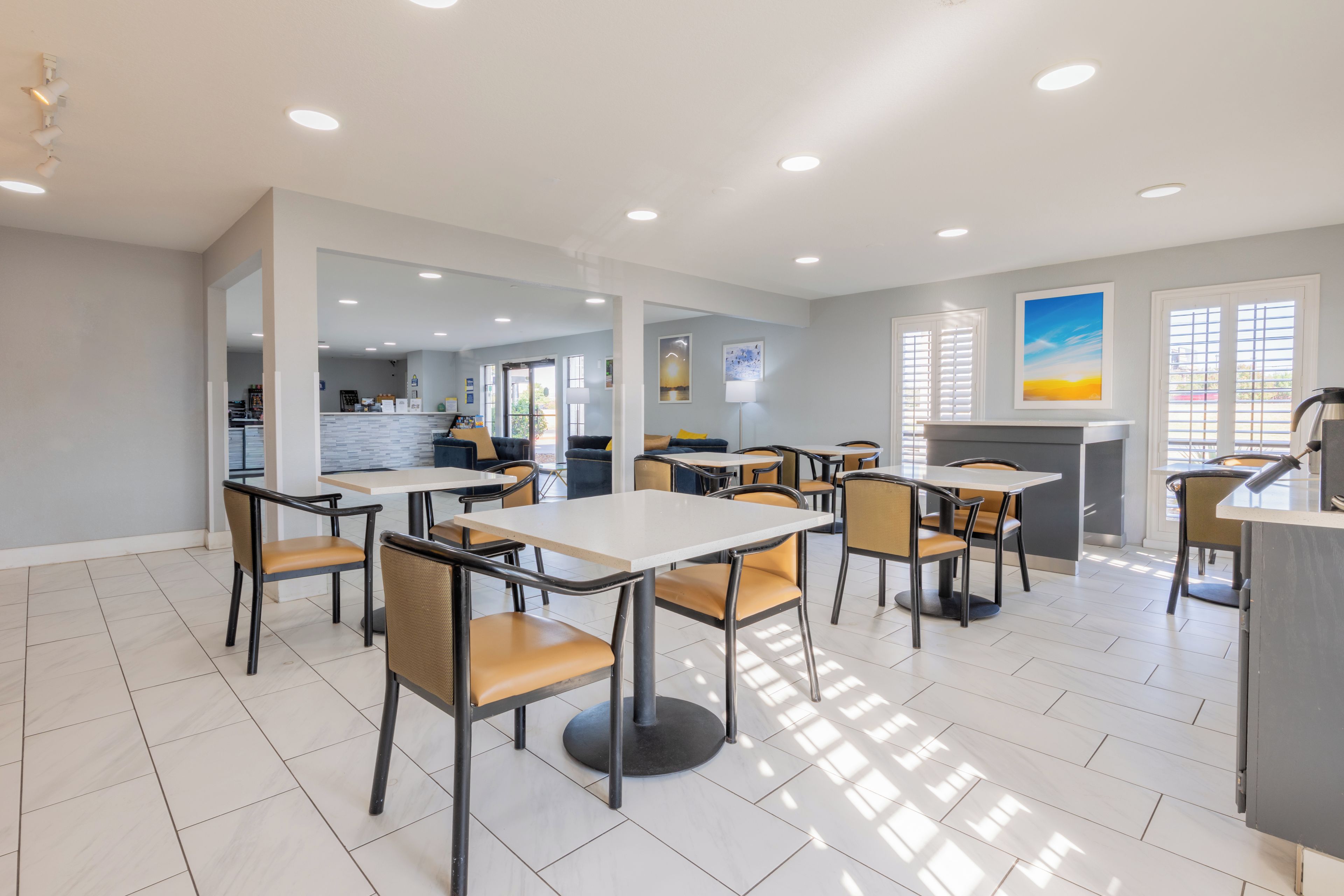 Un comedor moderno con mesas y sillas, una cocina al fondo y vistas al exterior a través de amplios ventanales.