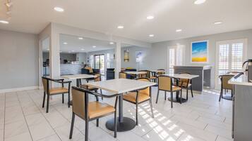 Un comedor moderno con mesas y sillas, una cocina al fondo y vistas al exterior a través de amplios ventanales.