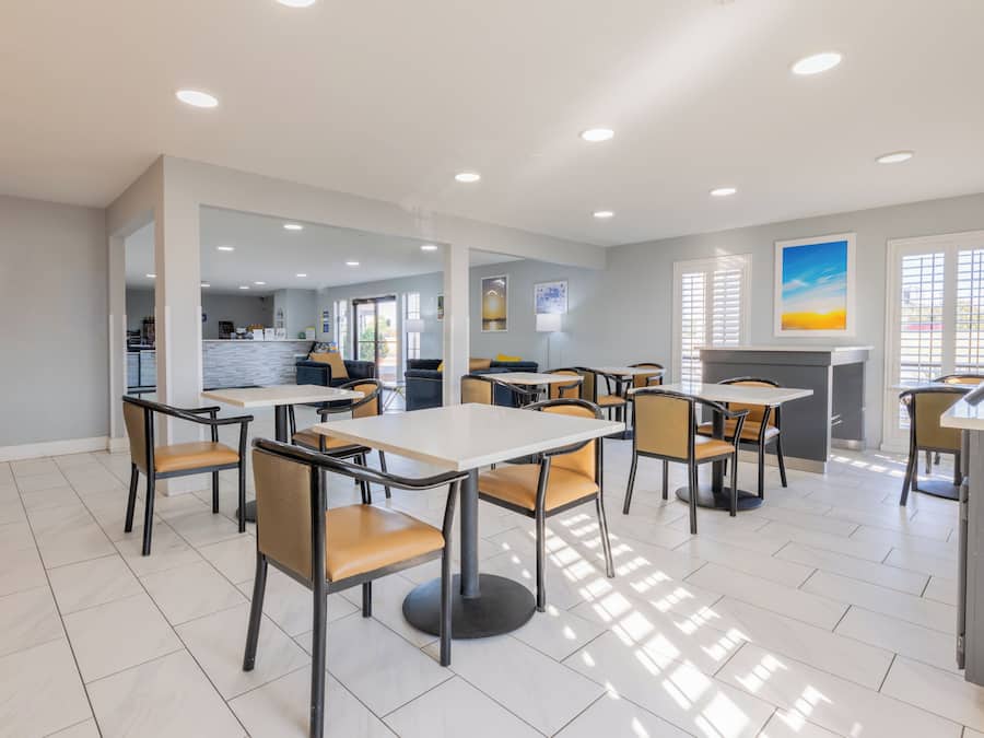 A modern dining area with tables and chairs, a kitchen in the background, and a view of the outdoors through large windows.