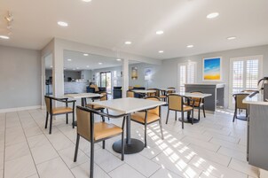 Un comedor moderno con mesas y sillas, una cocina al fondo y vistas al exterior a través de amplios ventanales.