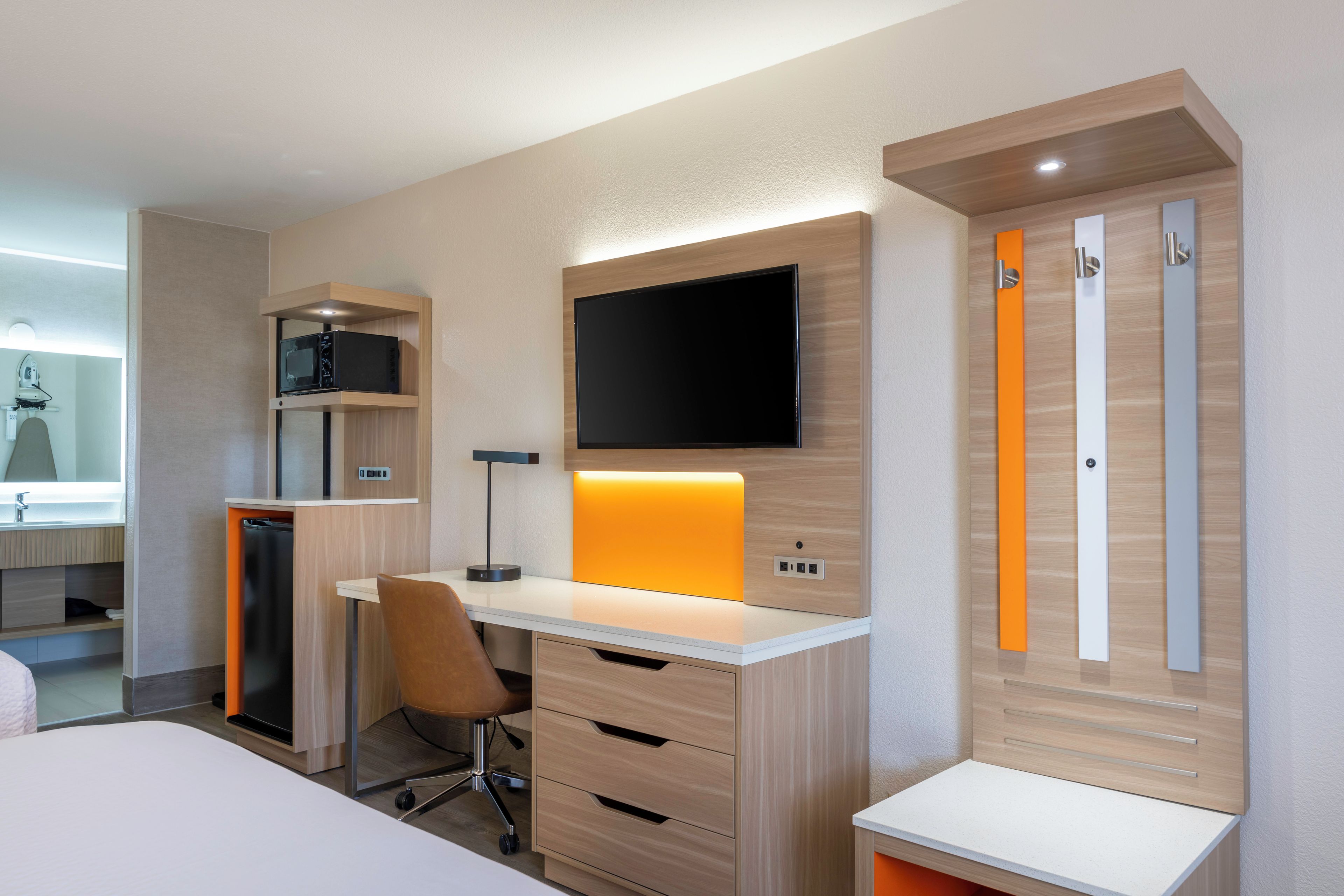A modern hotel room with a desk, chair, TV, wardrobe, and built-in appliances.