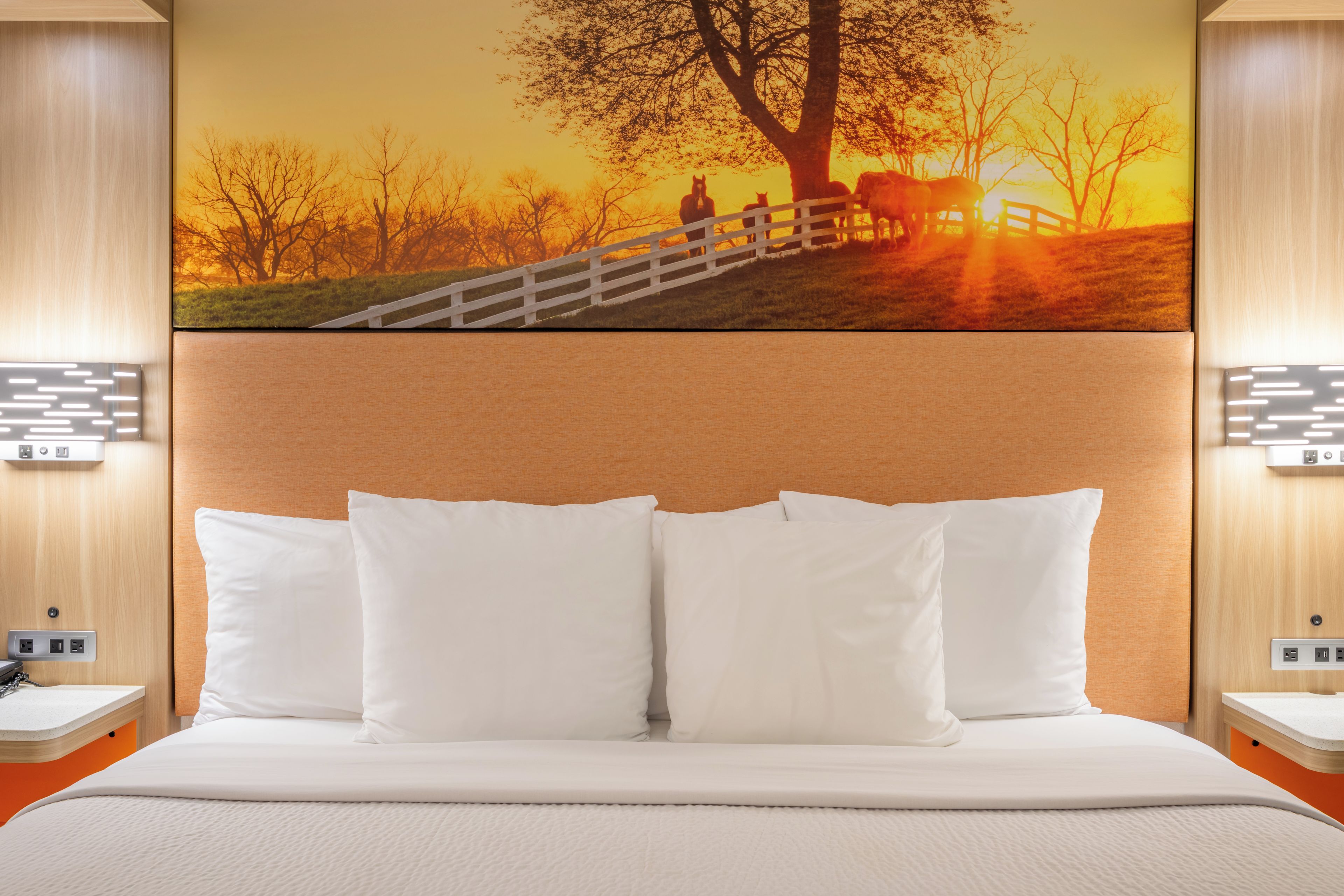 Habitación de hotel con una cama grande, ropa de cama blanca, cabecera con una pintura de atardecer y mesitas de noche con lámparas.
