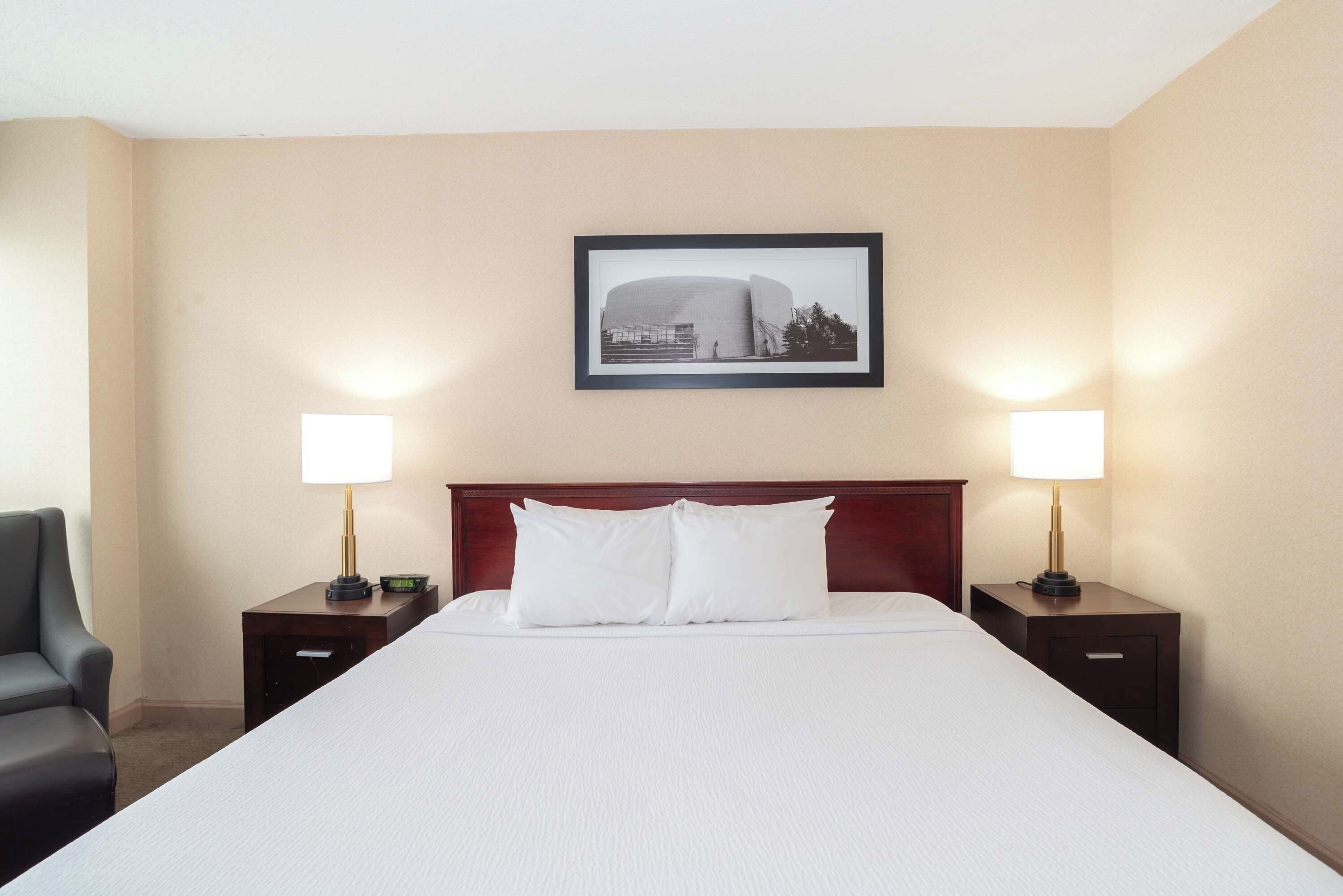 Ein Hotelzimmer mit einem großen Bett, zwei Nachttischlampen, einem gerahmten Bild an der Wand und einem Sessel.
