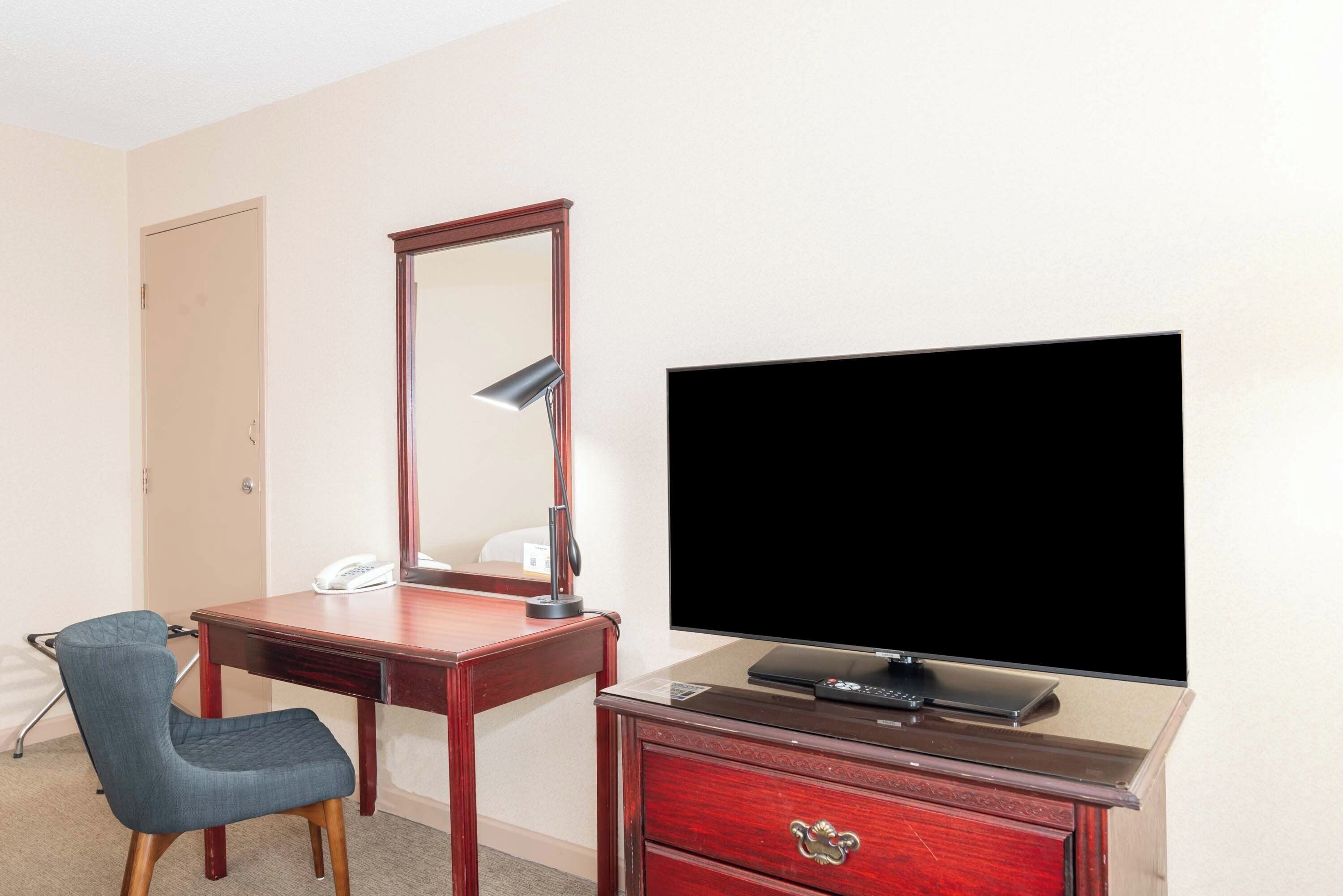 A hotel room with a wooden desk, a chair, a television, and a dresser.