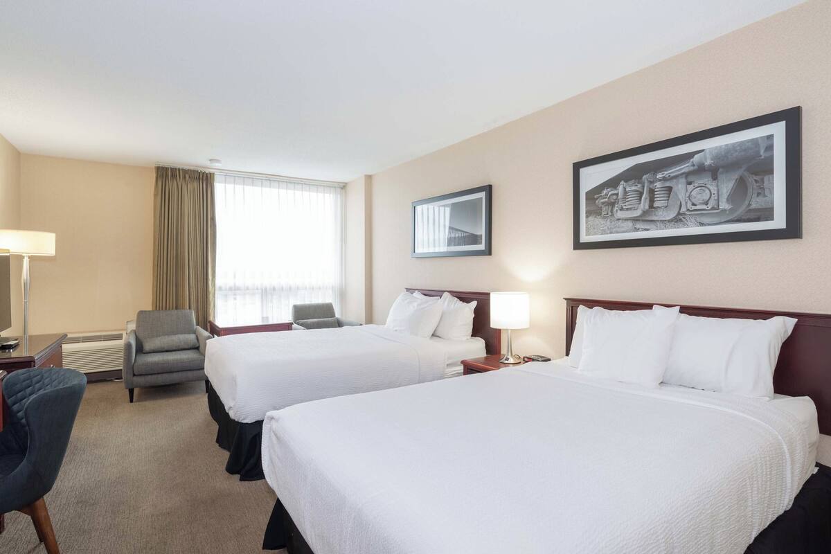 A hotel room with two beds, a desk, a chair, a lamp, and a window with curtains.