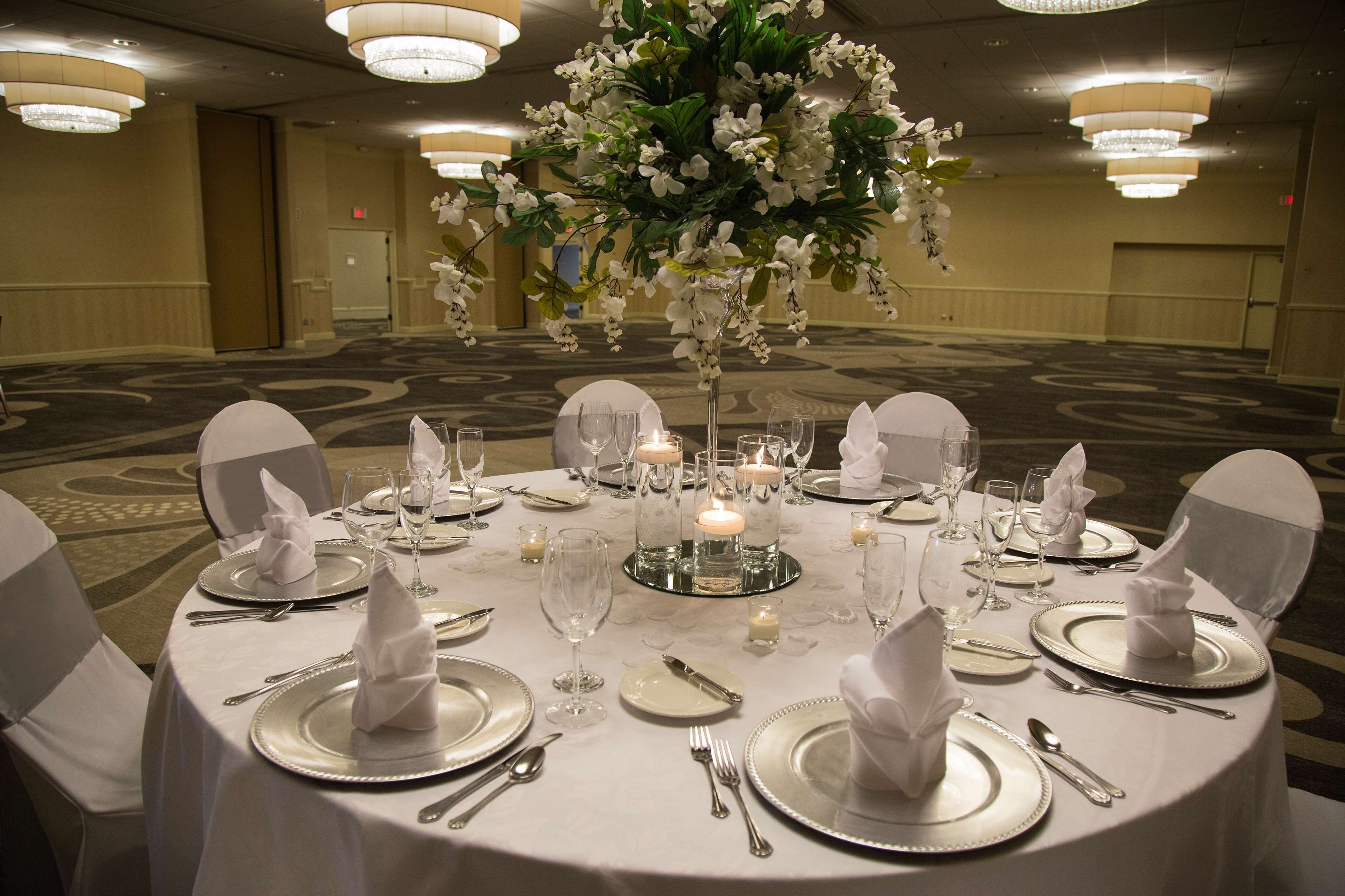 Una mesa redonda dispuesta para un evento formal, con manteles blancos, cubiertos y un centro de mesa floral.