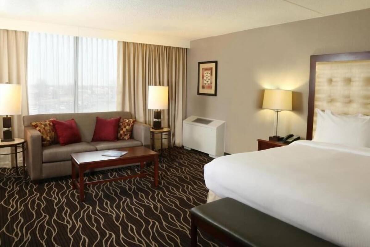 Una habitación de hotel con cama, sofá, mesa de centro, dos lámparas, ventana con cortinas y aire acondicionado instalado en la pared.