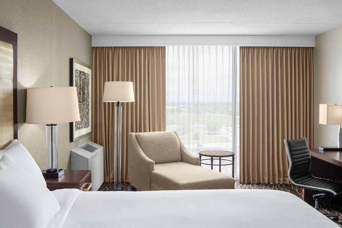Habitación de hotel con una cama grande, dos sillas, un escritorio, una lámpara y vistas a la ciudad desde la ventana.