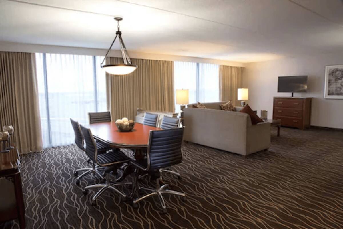 Habitación de hotel con zona de comedor, sofá, televisión y alfombra estampada.