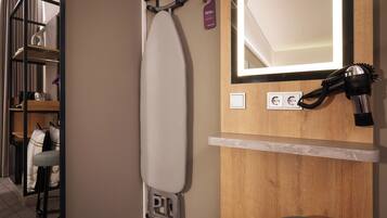 A hotel room with a wooden vanity, an ironing board, a hairdryer, and a mirror.