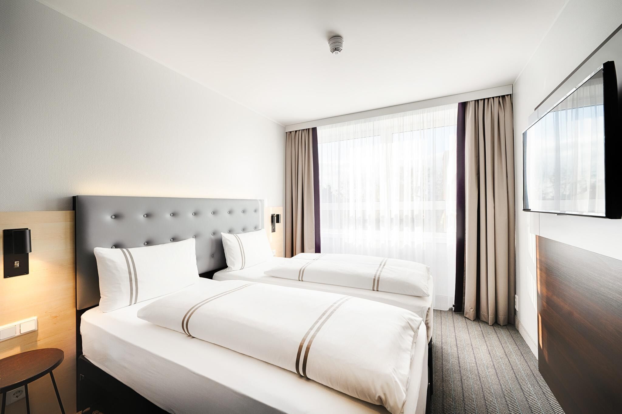 A hotel room with two beds, a flat-screen TV, a desk, and a window with curtains.