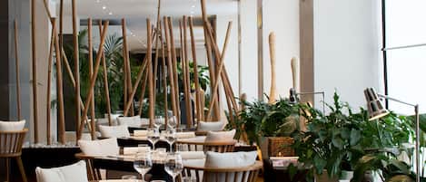 A modern dining area with wooden furniture, tables set with wine glasses and candles, and a large window providing natural light.
