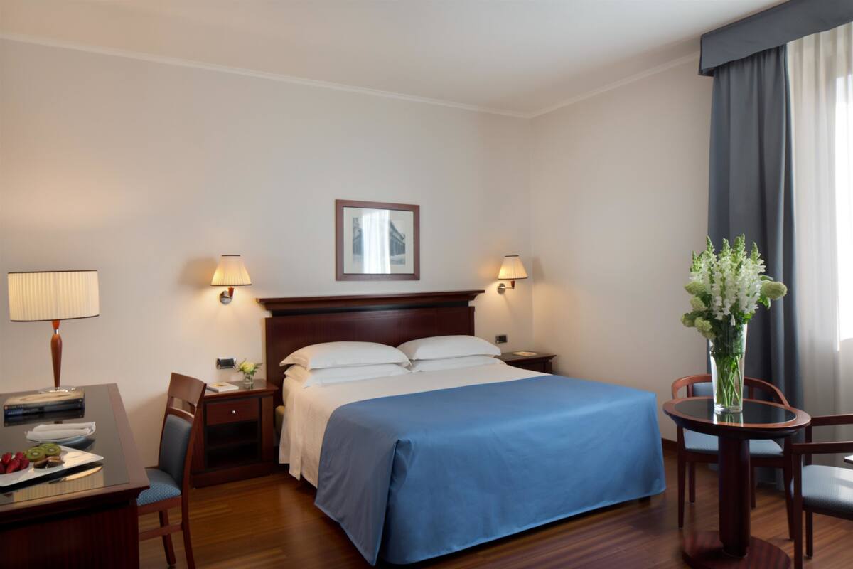A hotel room with a bed, bedside tables, a desk with a lamp, a chair, and a vase with flowers.