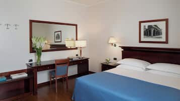Habitación de hotel con cama, escritorio, silla, mesita de noche, lámpara, espejo y un cuadro en la pared.