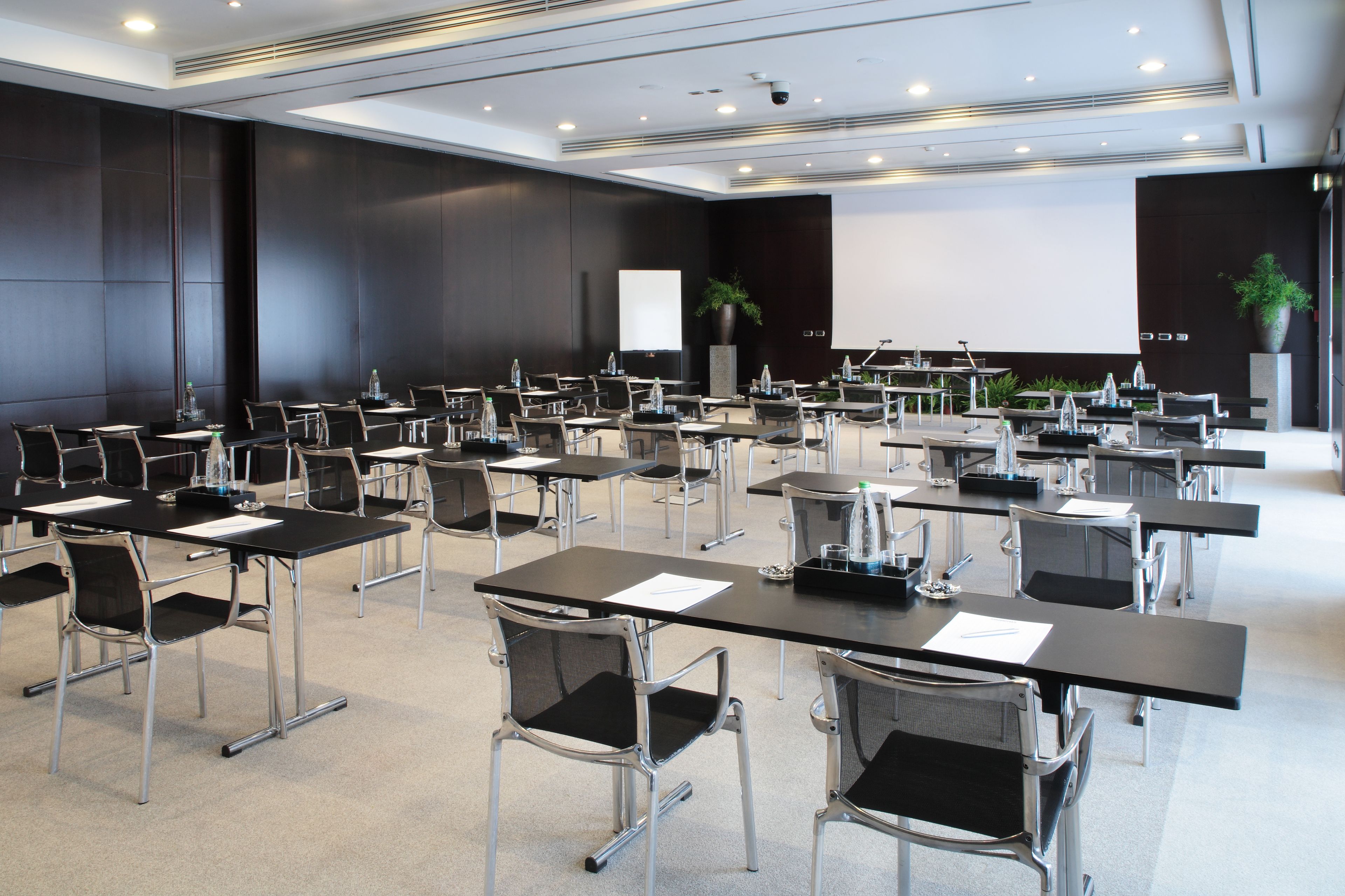 Une salle de conférence équipée de plusieurs tables et chaises, d’un écran de projection et d’une scène avec un pupitre.