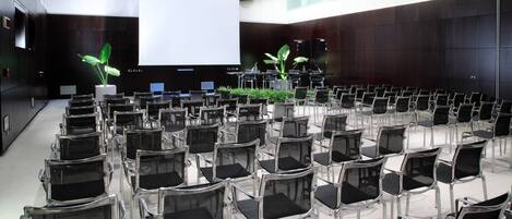 A conference room with rows of chairs, a large screen, and two potted plants.
