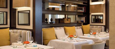 A restaurant with multiple tables, each set with wine glasses, plates, and cutlery. There are cushioned chairs and a sofa with striped cushions.