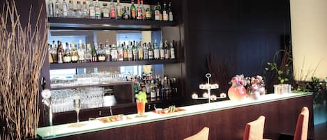 Zona de bar con dos estantes llenos de botellas de licor, una barra y cuatro sillas con cojines.