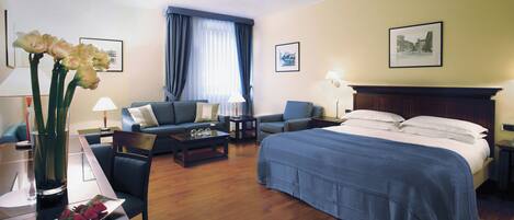 Une chambre d’hôtel comprenant un lit, deux fauteuils, une table basse, un bureau et un vase avec des fleurs.