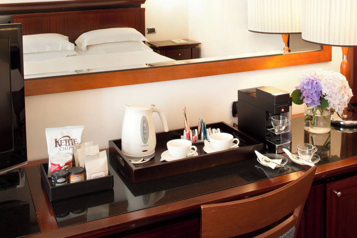 A hotel room with a bed, a bedside table with a tray containing a coffee maker, cups, and snacks, and a mirror above the bed.