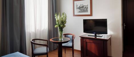 A hotel room with a bed, a TV on a wooden cabinet, a small round table with a vase of flowers, two chairs, and a framed picture on the wall.