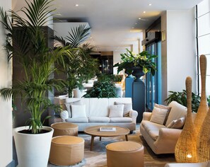 A modern living room with a sofa, ottomans, a coffee table, and large potted plants. - Starhotels Excelsior (Bologna)
