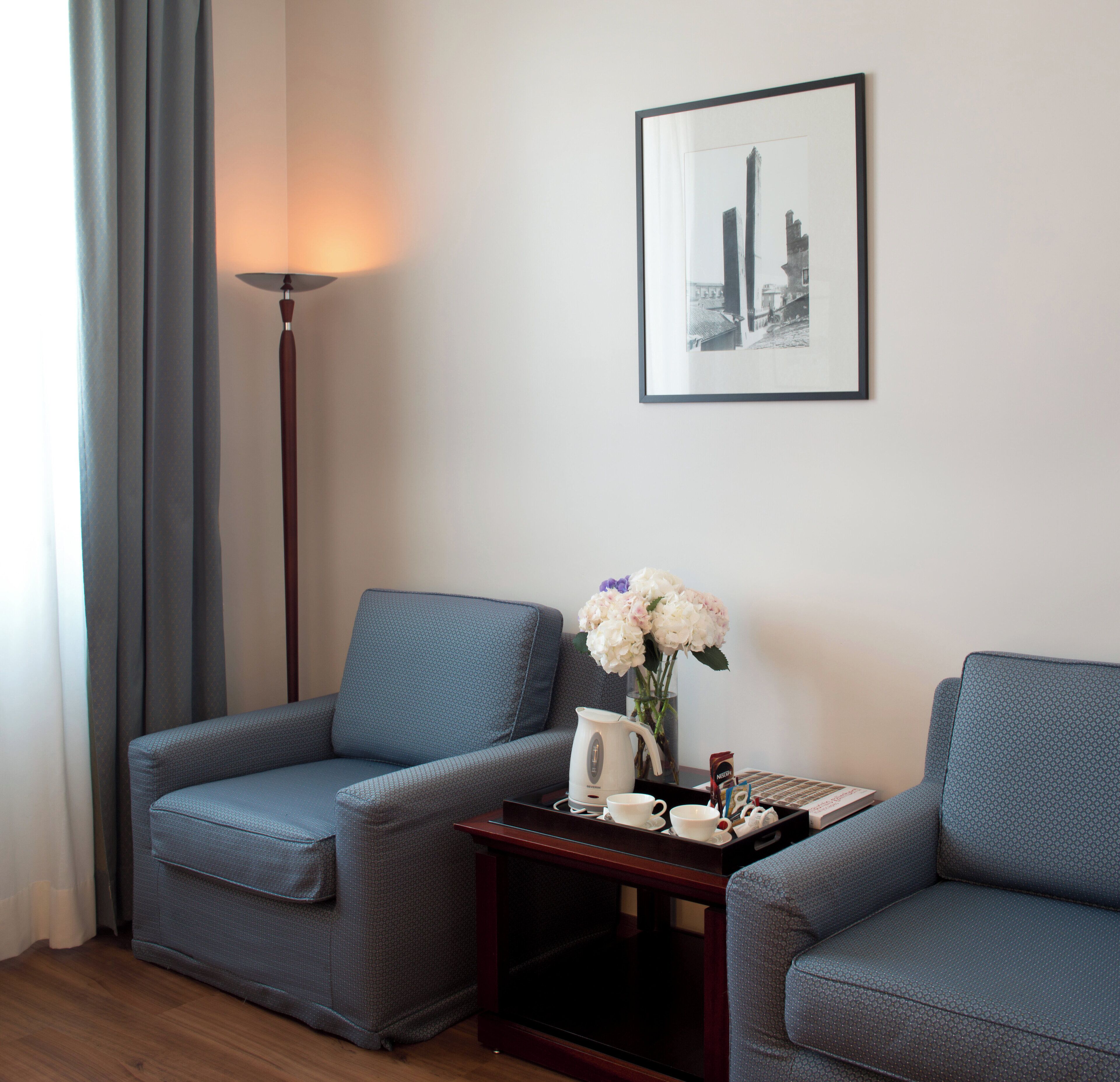 A room with two grey sofas, a small wooden side table with a vase of flowers, a framed picture on the wall, and a floor lamp.
