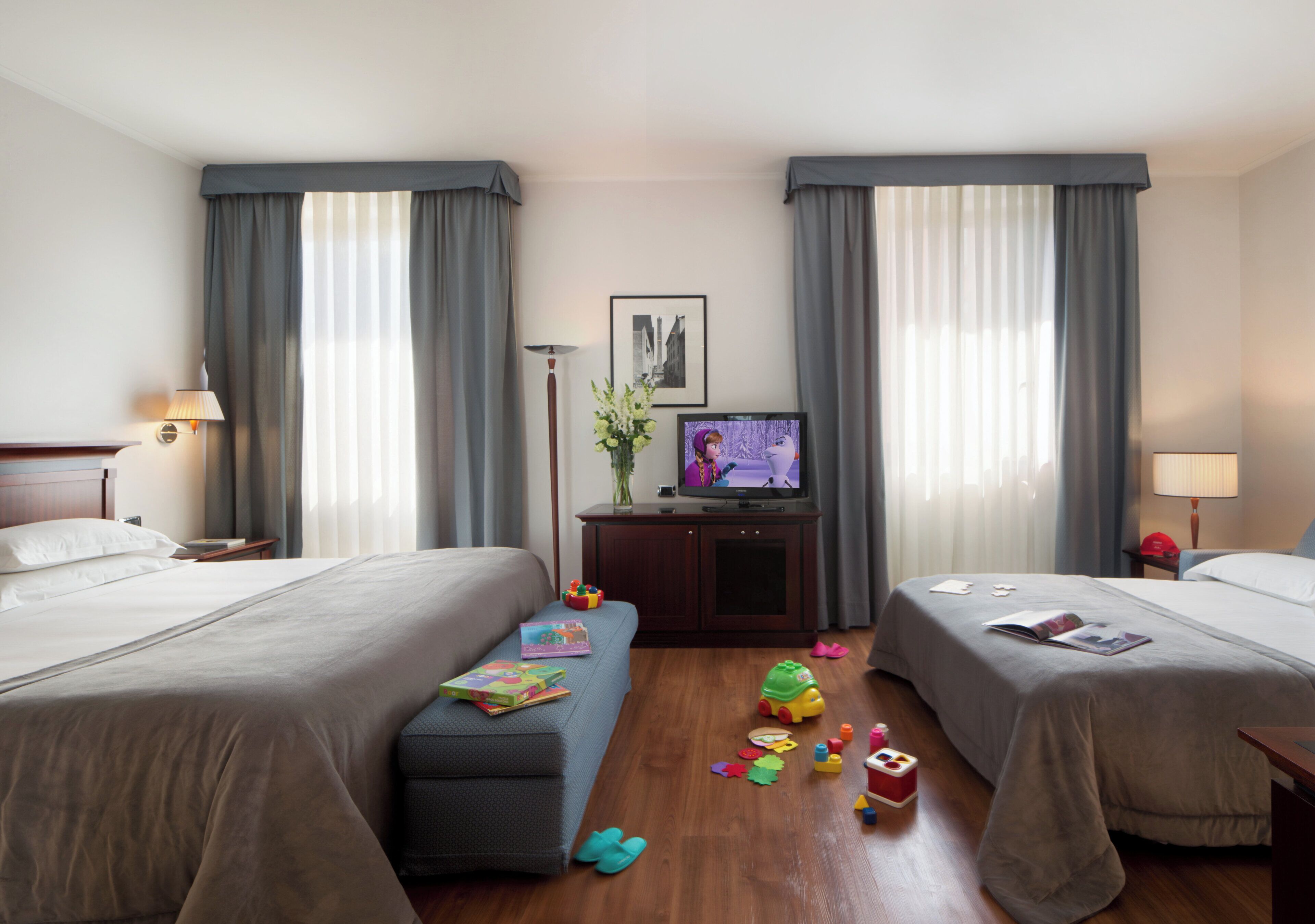 A hotel room with two beds, a TV, a desk with a lamp, and toys on the floor.