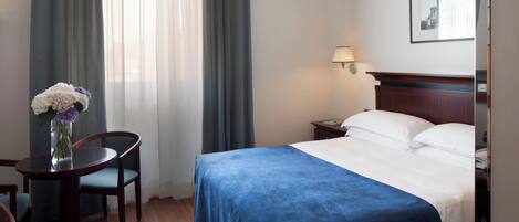 Quarto de hotel com cama, escrivaninha com cadeira, um vaso de flores e uma luminária.