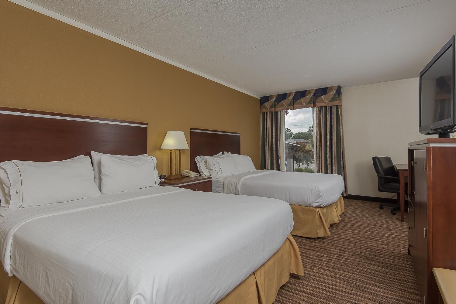 A hotel room with two beds, a TV, a desk, and a window with curtains.