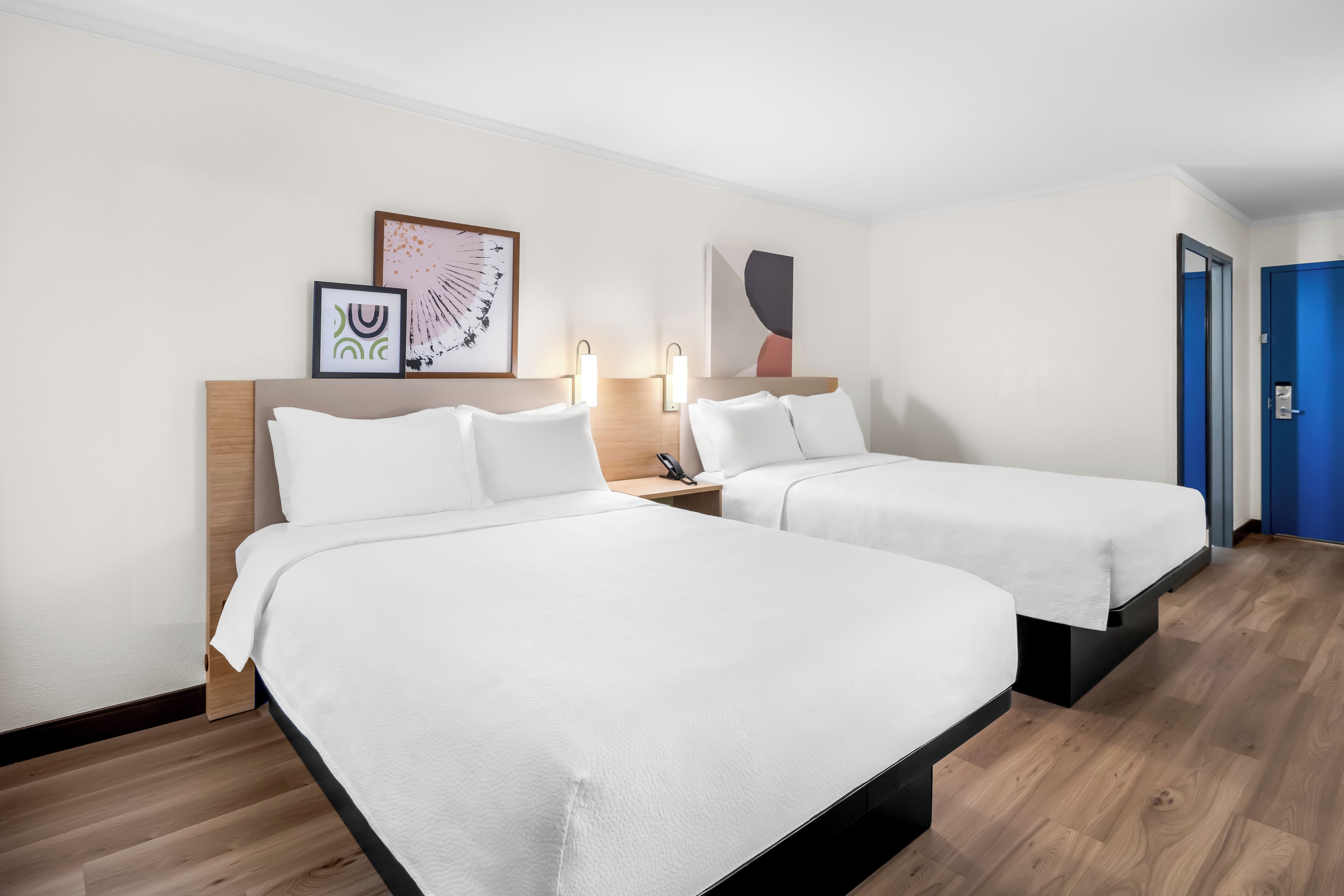 A hotel room with two single beds, a wooden headboard, bedside tables with lamps, and artwork on the walls.