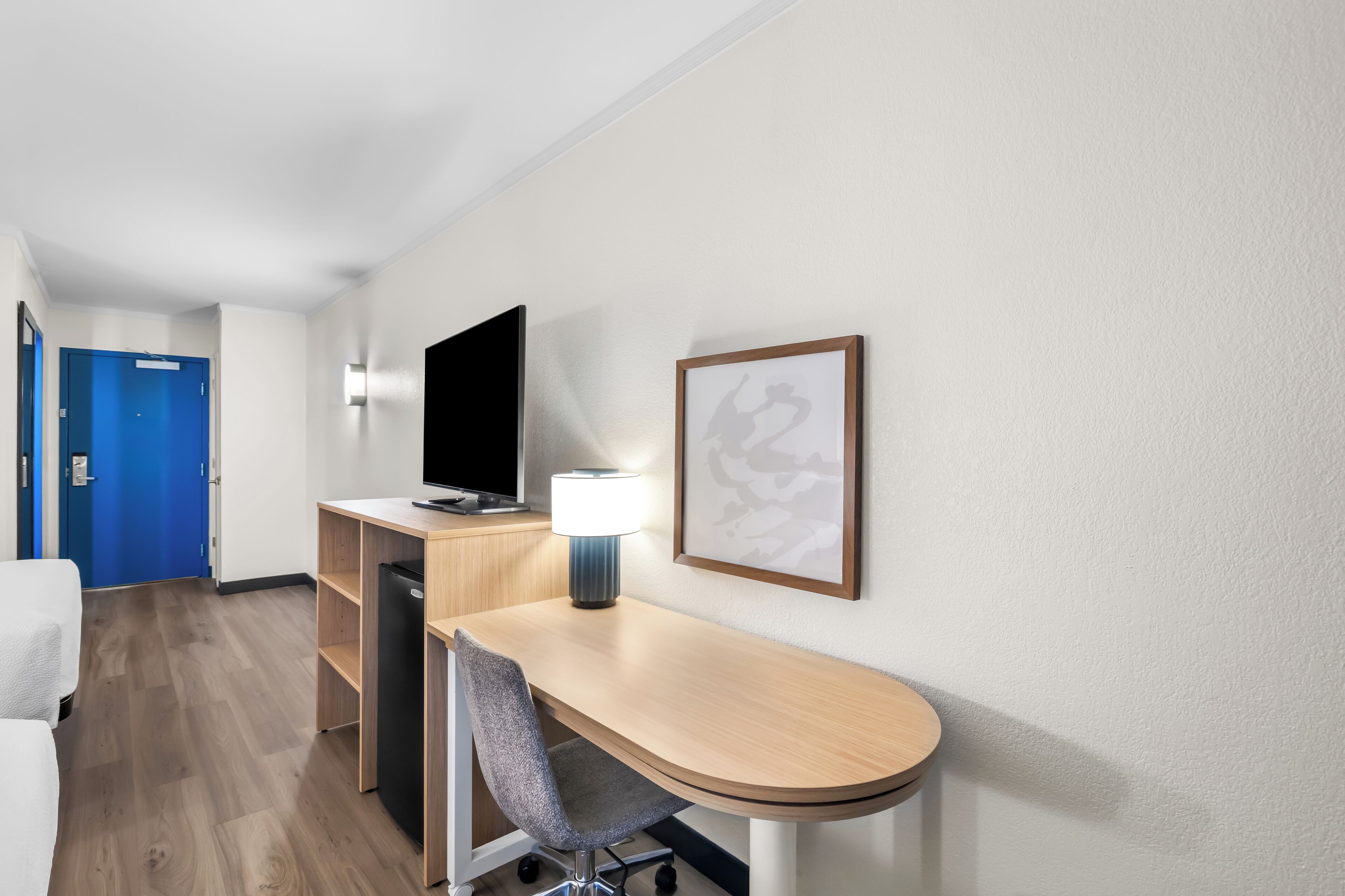 A hotel room with a desk, chair, TV, lamp, and artwork on the wall.