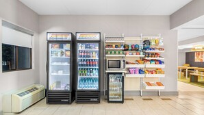 A clean, modern food service area with three beverage dispensers, a microwave, and shelves stocked with snacks and beverages. - Spark by Hilton Roanoke Civic Center (Roanoke)