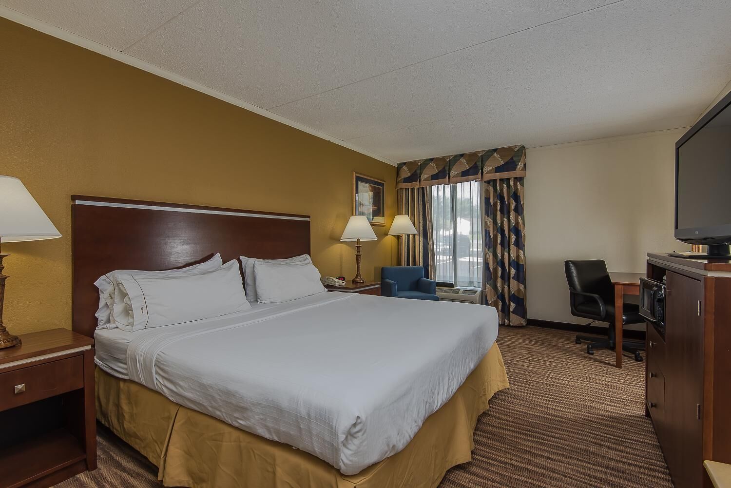 A hotel room with a large bed, two bedside lamps, a desk with a chair, a television, and a window with curtains.