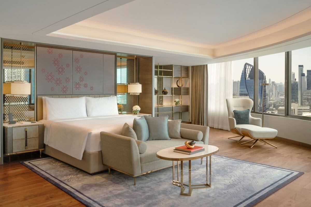 A modern hotel room with a large bed, a sitting area with a sofa and armchair, a desk, and a view of the city skyline.