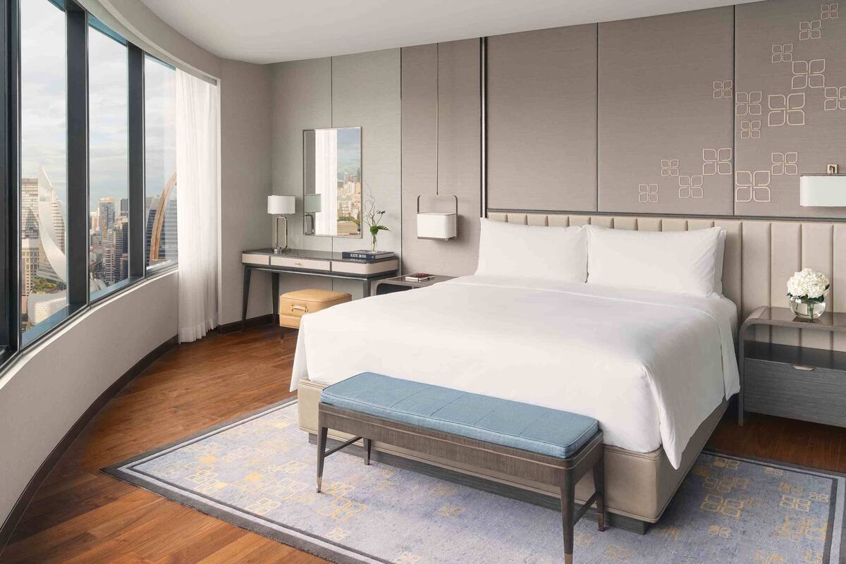 A modern hotel room with a large bed, a desk, a chair, a bench, and a view of the city through large windows.