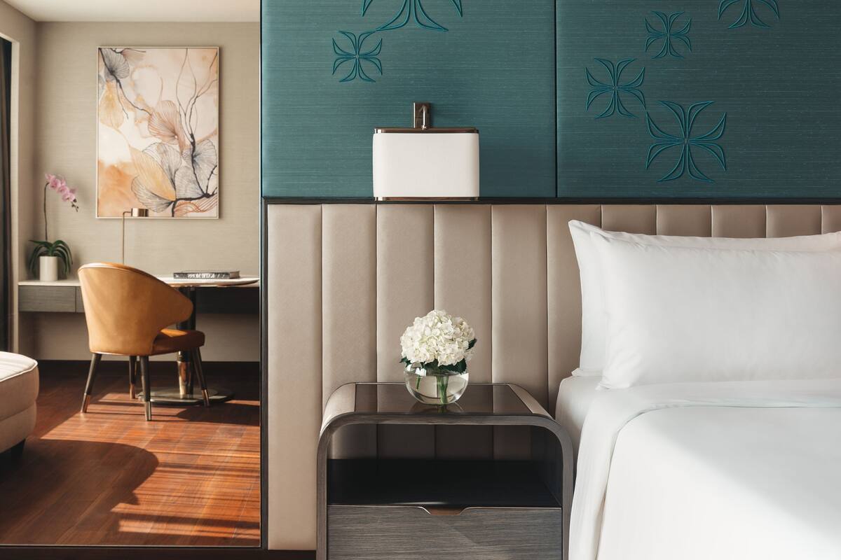 A modern hotel room with a bed, a nightstand with a vase of flowers, a chair, a desk, and a large abstract painting on the wall.