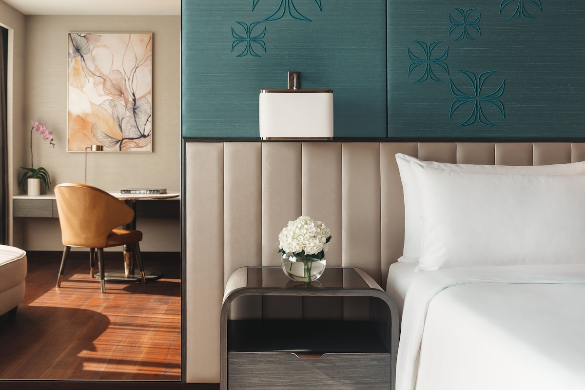 A modern hotel room with a bed, a nightstand with a vase of flowers, a chair, a desk, and a large abstract painting on the wall.