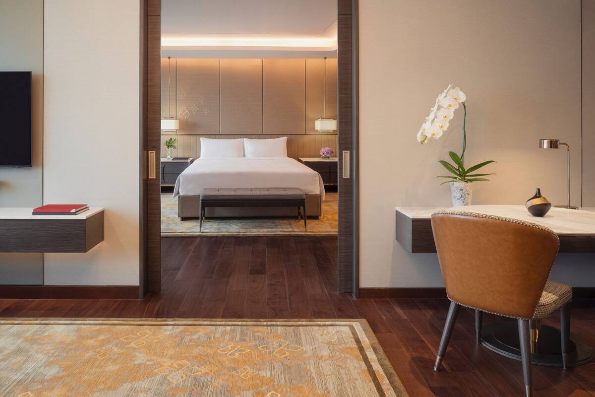 A modern hotel room with a large bed, a desk with a chair, a TV, and a plant.