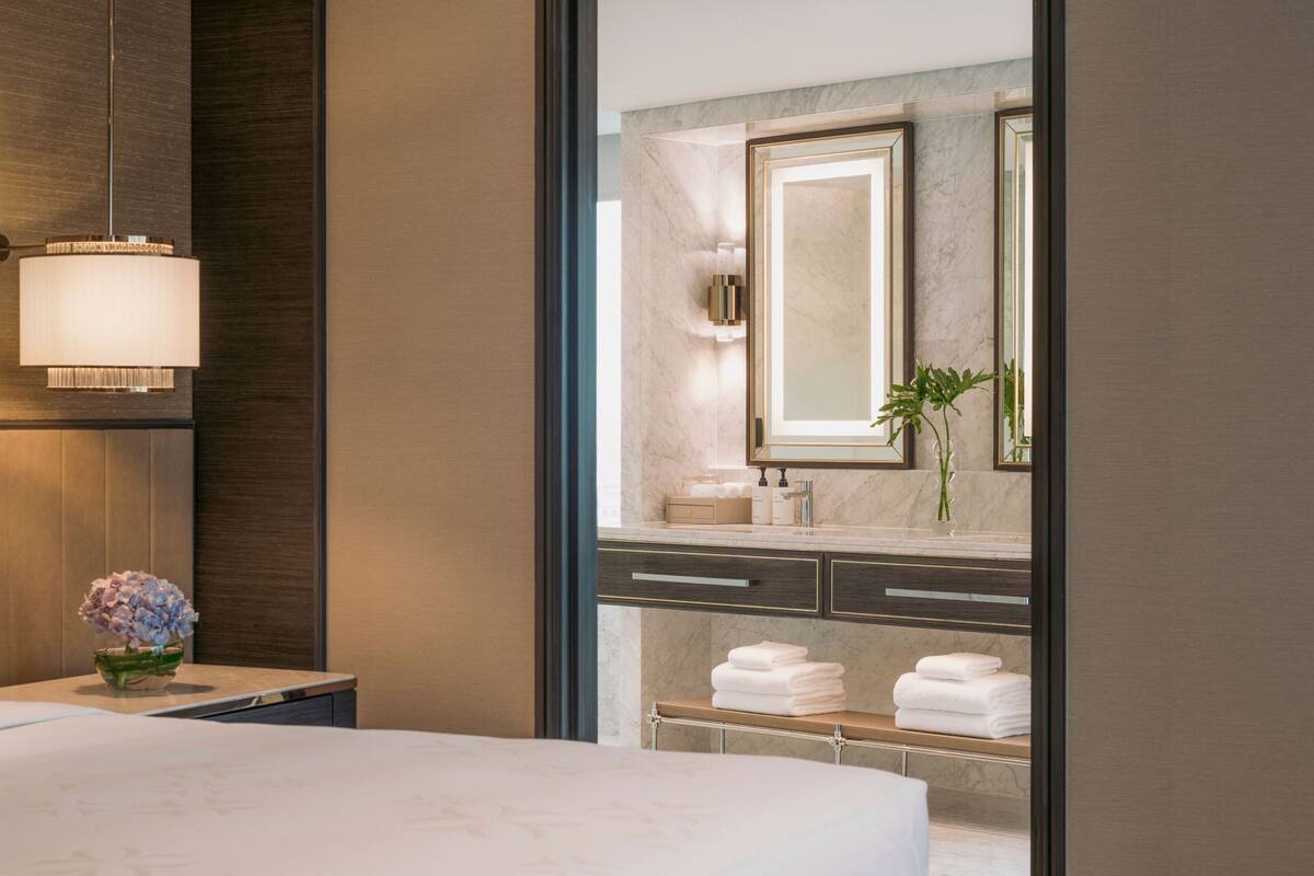 A hotel room with a large bed, a bedside table with a lamp, a nightstand with a vase of flowers, and a view of the bathroom through a glass door.