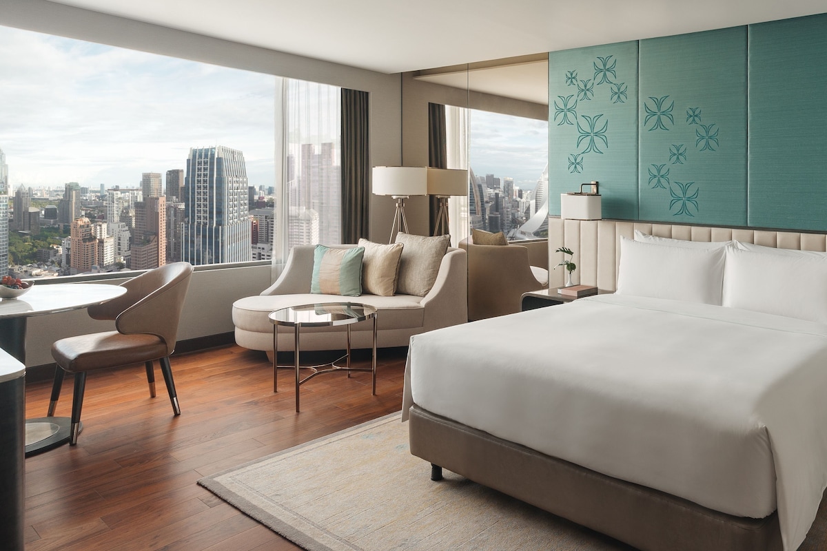 A modern hotel room with a large bed, a sofa, a desk, and a view of the city skyline.
