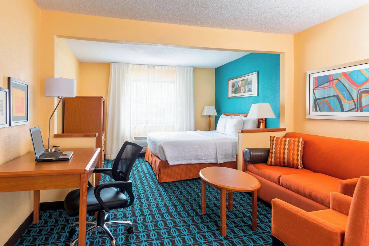 A hotel room with a bed, a desk with a laptop, a sofa, a chair, and a small table.